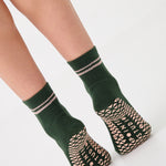 Woman wearing a solid green ankle grip sock with pink stripes on the toes and top ankle cuff made for Pilates, Barre, Yoga, and beyond. 