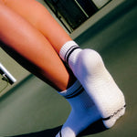 Women wearing a white ankle grip sock with black stripes on the toes and top ankle cuff made for Pilates, Barre, Yoga, and beyond. 