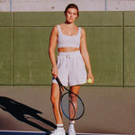Women wearing a white ankle grip sock with black stripes on the toes and top ankle cuff made for Pilates, Barre, Yoga, and beyond. She's at a tennis court ready to play. 