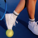 Women wearing a white ankle grip sock with black stripes on the toes and top ankle cuff made for Pilates, Barre, Yoga, and beyond. She's at a tennis court ready to play.