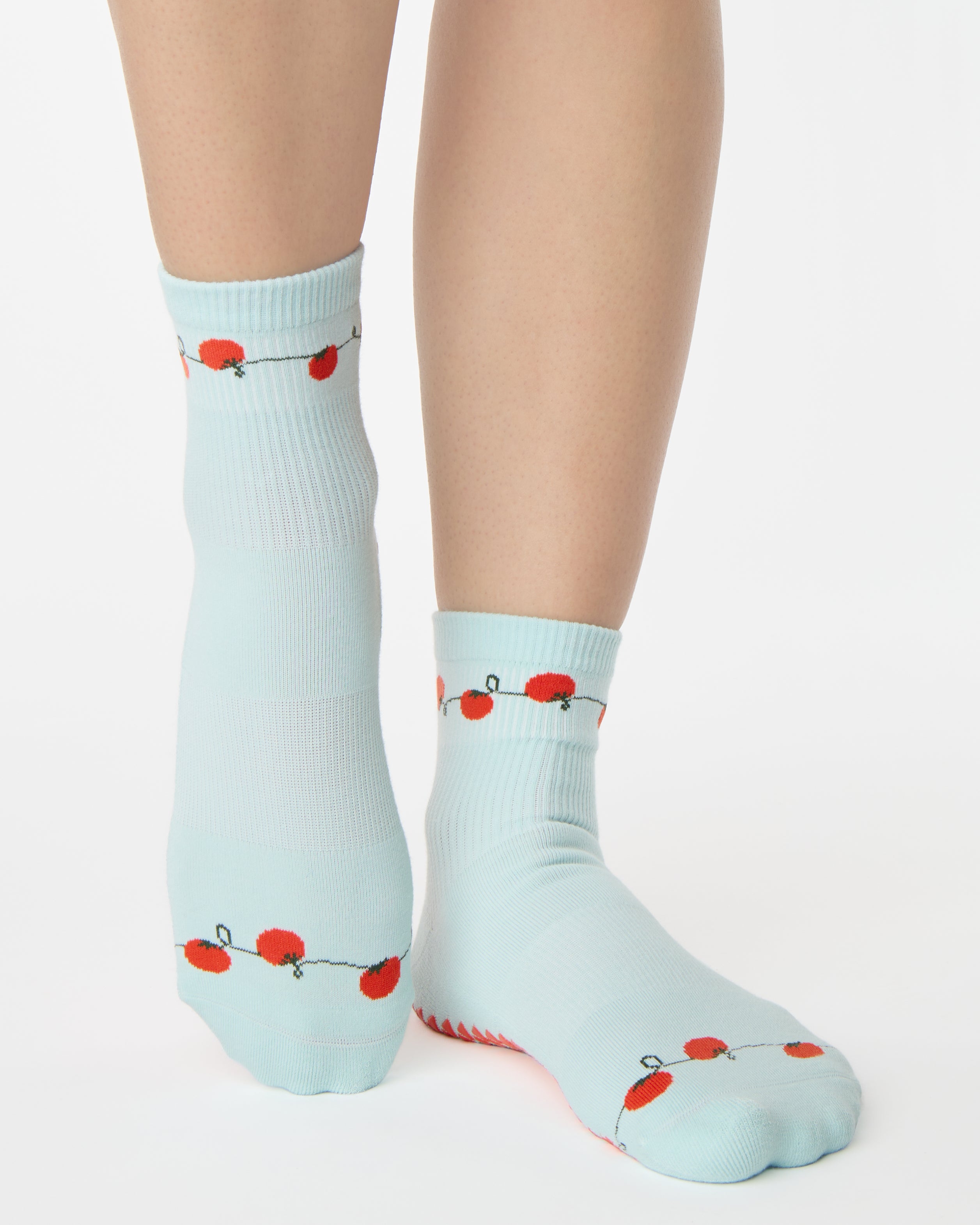 Woman wearing a solid light blue ankle grip sock with red tomatos on garden vines. Made for pilates, yoga, barre, and beyond. 