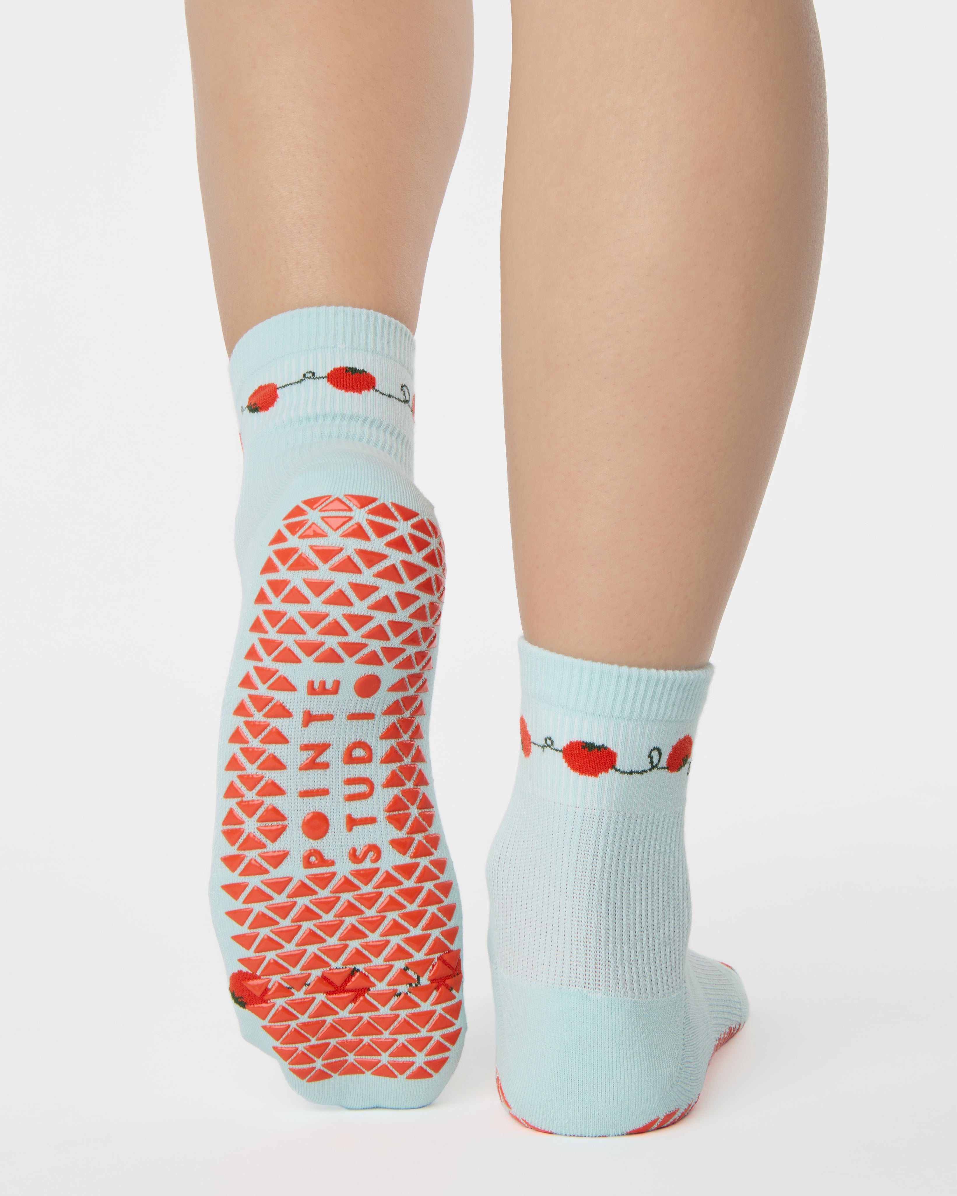 Woman wearing a solid light blue ankle grip sock with red tomatos on garden vines. Made for pilates, yoga, barre, and beyond. 