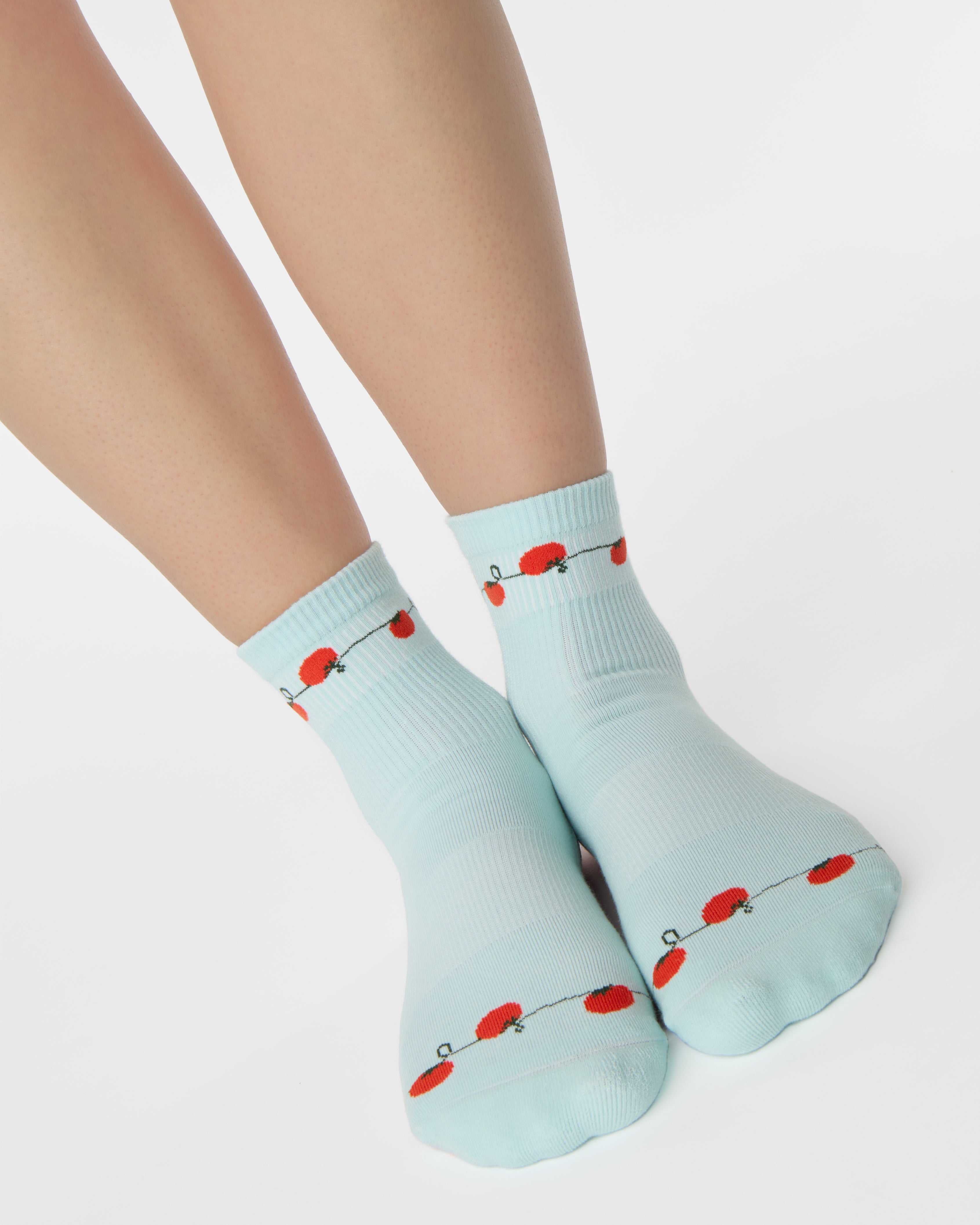 Woman wearing a solid light blue ankle grip sock with red tomatos on garden vines. Made for pilates, yoga, barre, and beyond. 