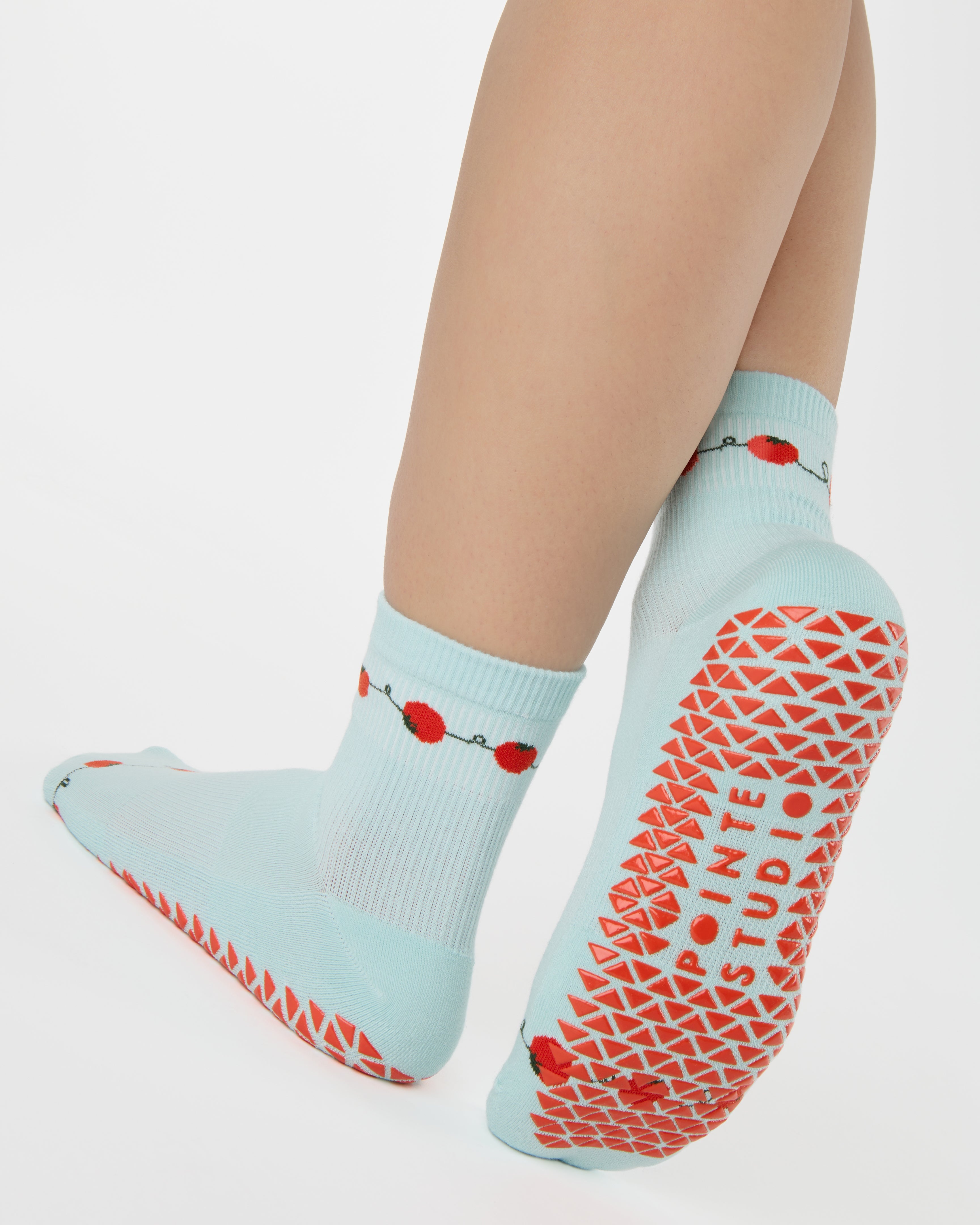 Woman wearing a solid light blue ankle grip sock with red tomatos on garden vines. Made for pilates, yoga, barre, and beyond. 