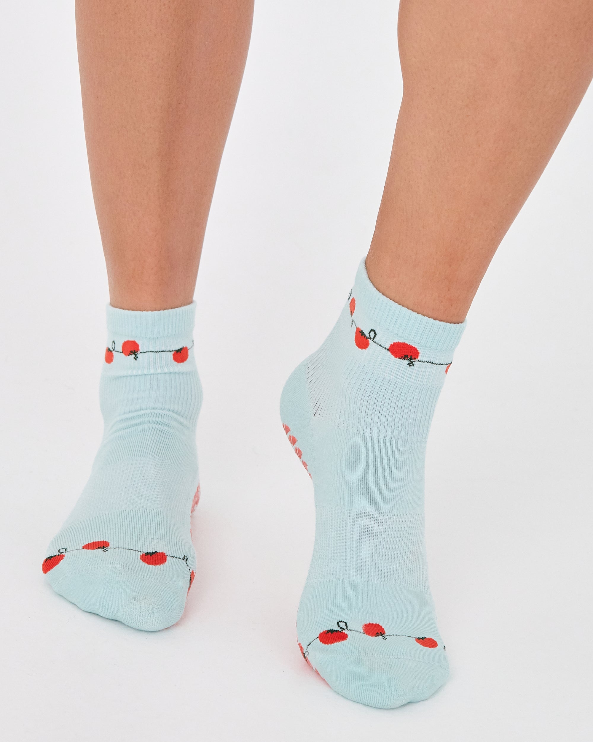 Woman wearing a solid light blue ankle grip sock with red tomatos on garden vines. Made for pilates, yoga, barre, and beyond. 